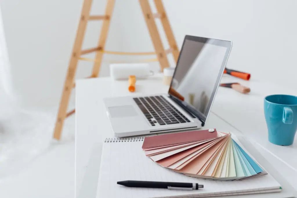 A DIY home remodel with a laptop, paint roller, coffee mug and colour samples on a table